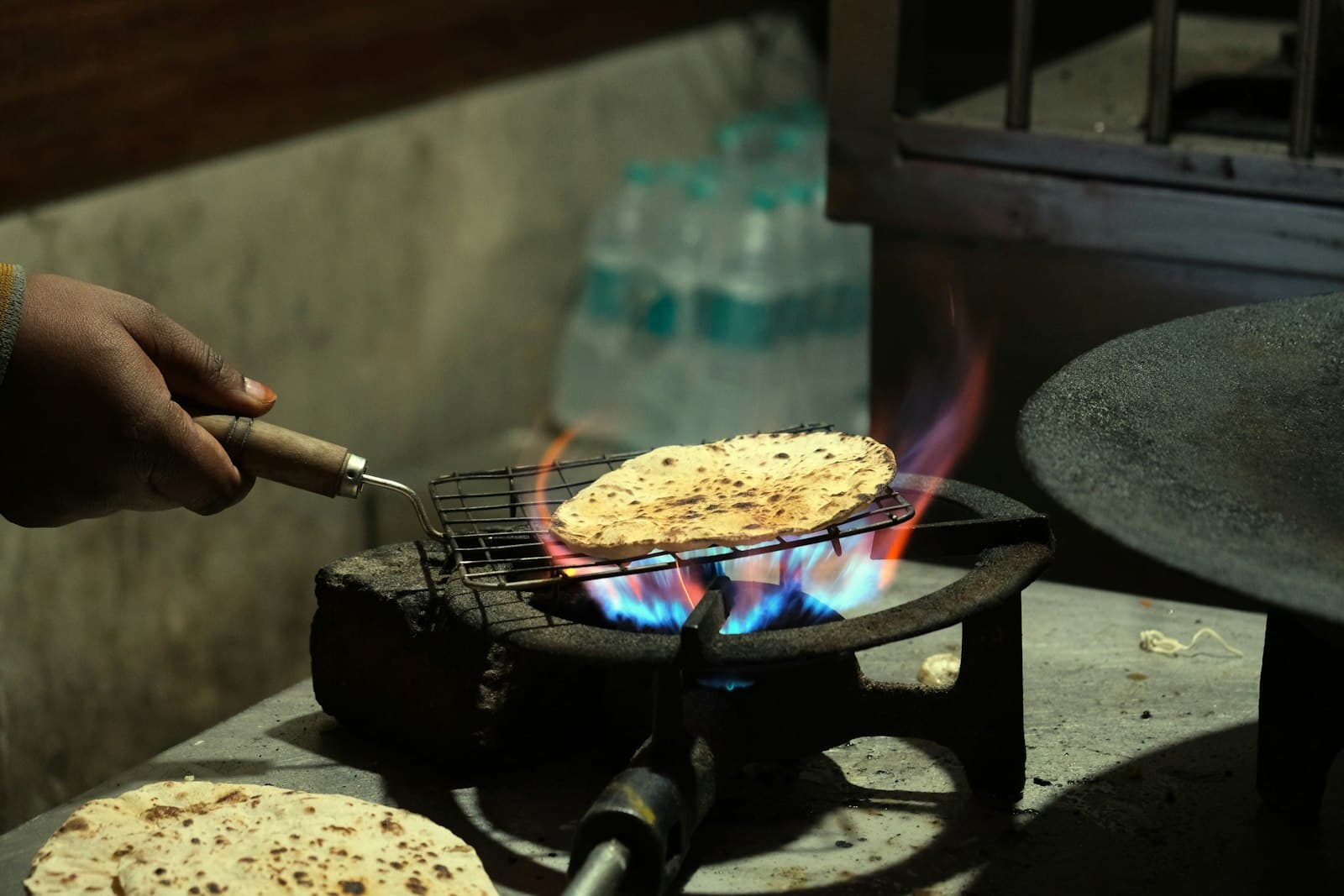 A hand grilling a roti directly on a blue gas flame — the daily action of an Indian home kitchen.