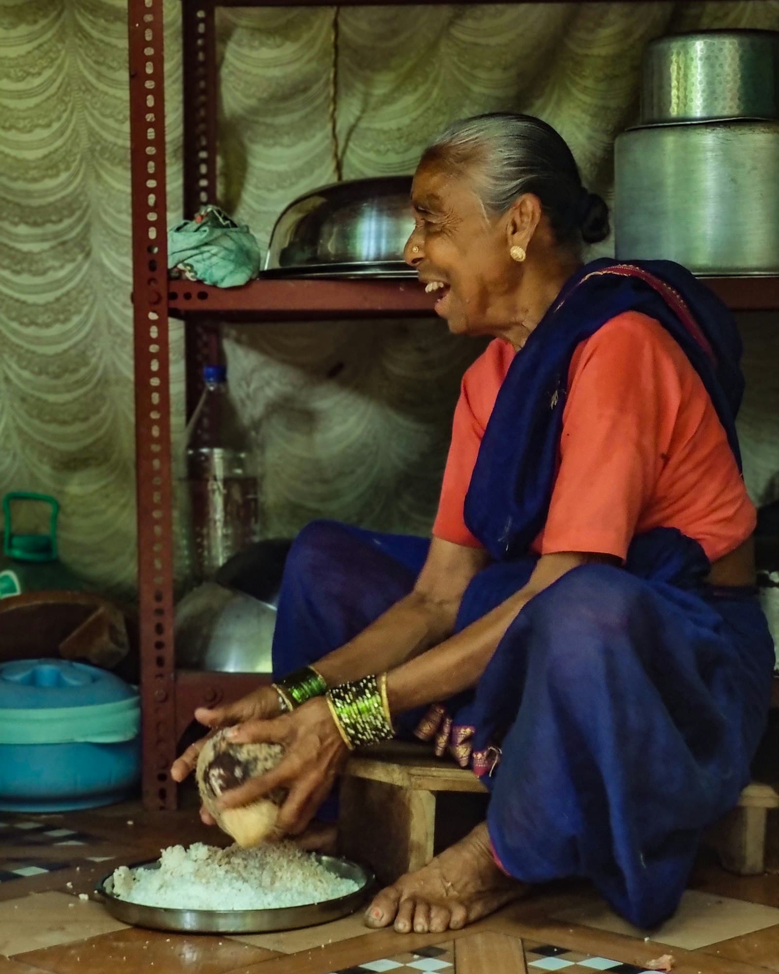A Konkani grandmother in a traditional Indian kitchen, preparing food with quiet care — the warmth of a household cook.