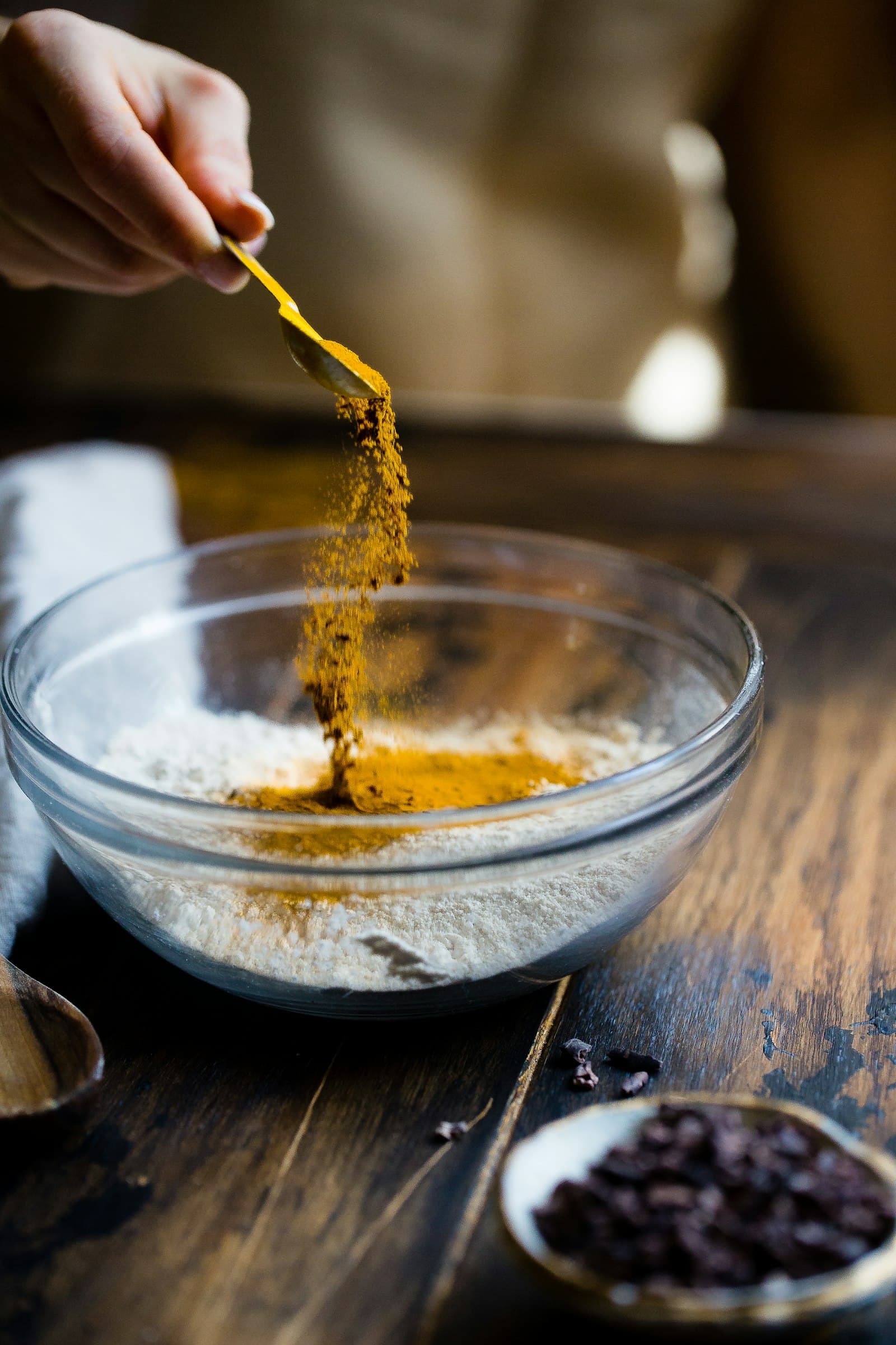 A hand pouring vibrant yellow turmeric from a wooden spoon into a small glass bowl — a measured cooking moment.