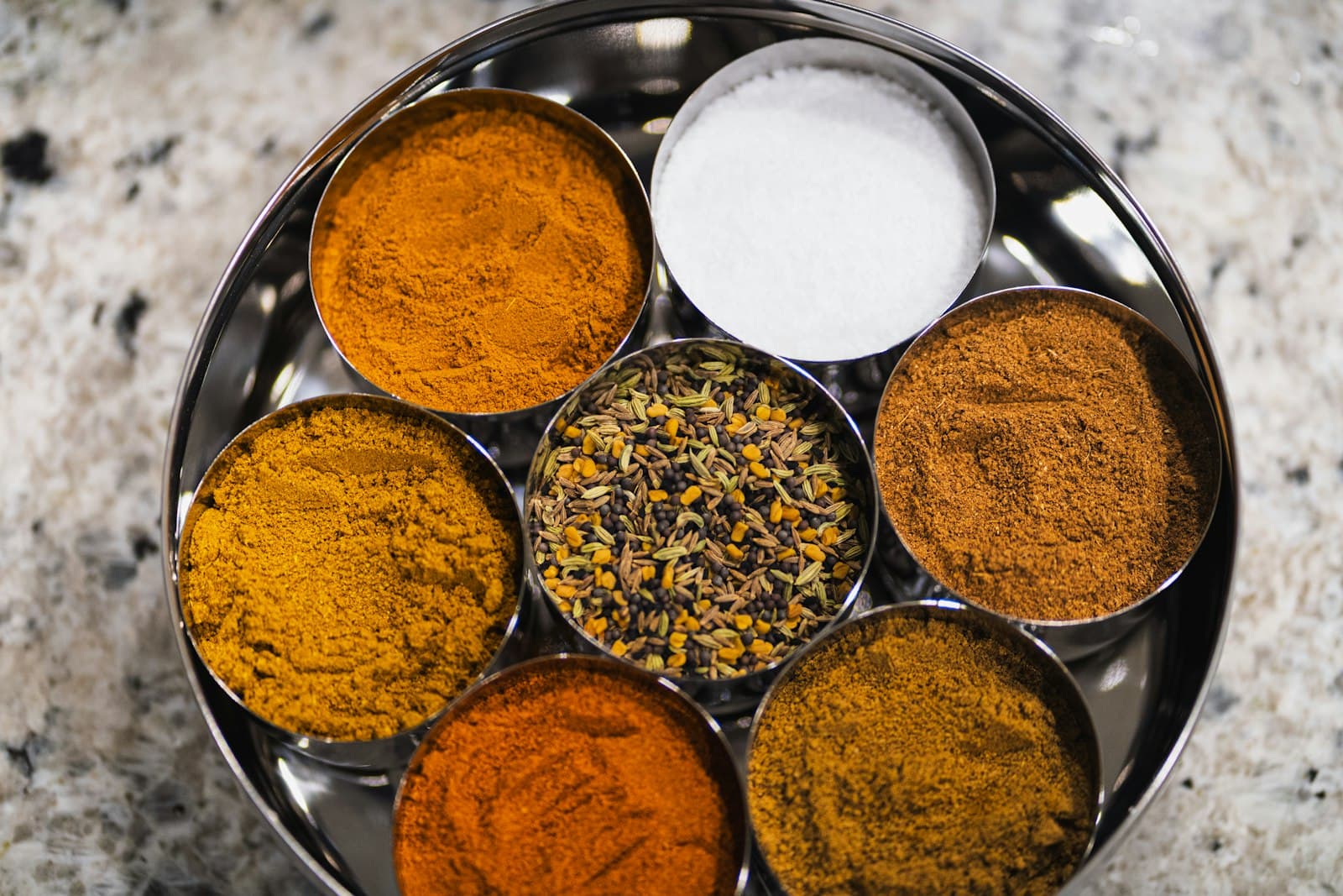 Colorful Indian spices arranged in metal bowls on a tray — turmeric, cumin, coriander, mustard.