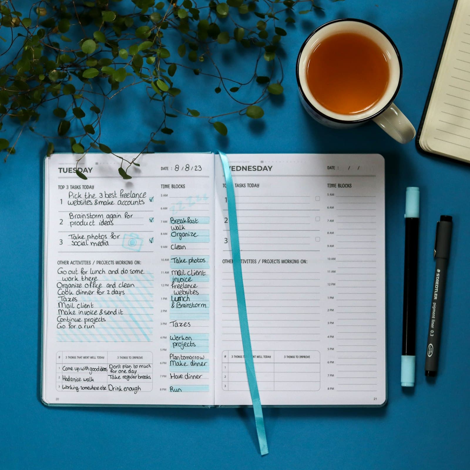 An open notebook on a wooden surface with a pen resting on the page and a small cup of tea beside it — the planner's flatlay.